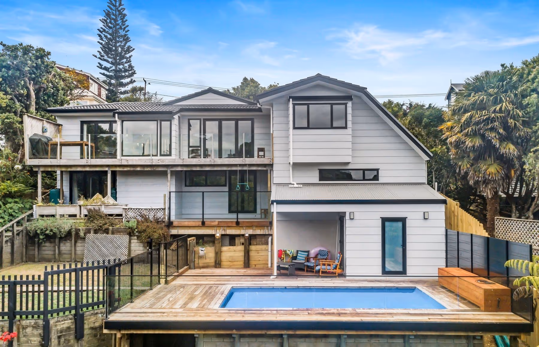 A modern two-story house with large windows, a wooden deck, and a rectangular pool, surrounded by lush greenery and a black fence.