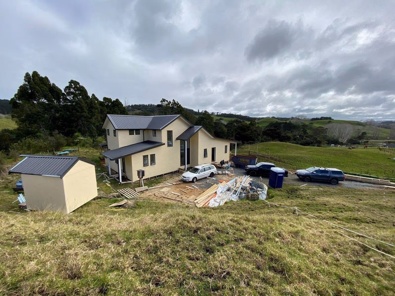 Modern two-story house under construction, with a gray metal roof, surrounded by building materials and a parked car on a grassy lot.