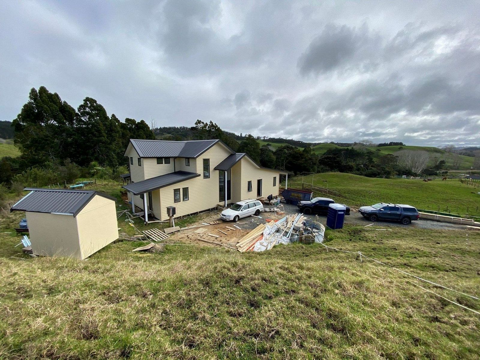 Modern two-story house under construction, with a gray metal roof, surrounded by building materials and a parked car on a grassy lot.
