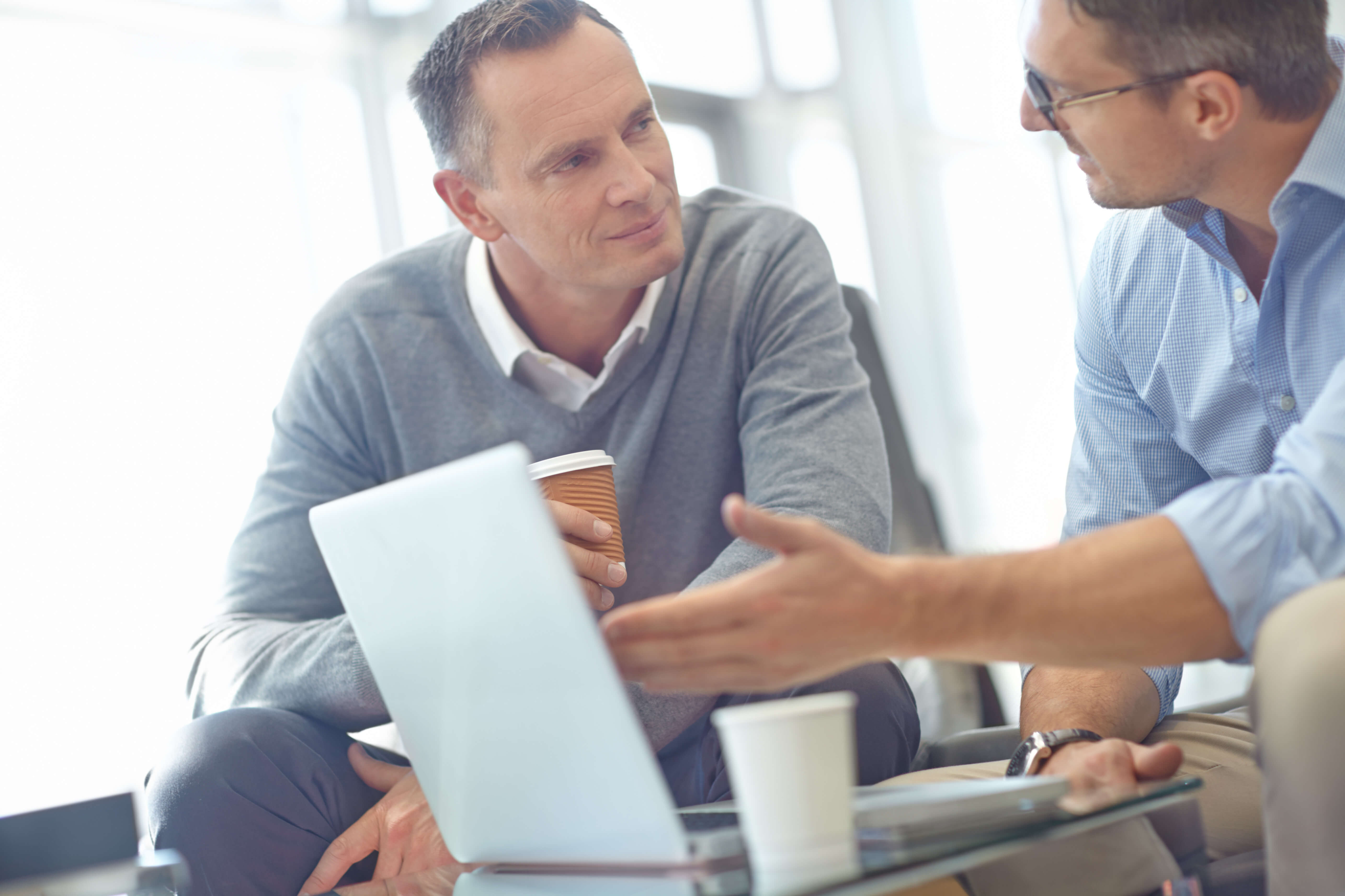 Two men sitting and talking while looking at a laptop, with coffee cups on the table.