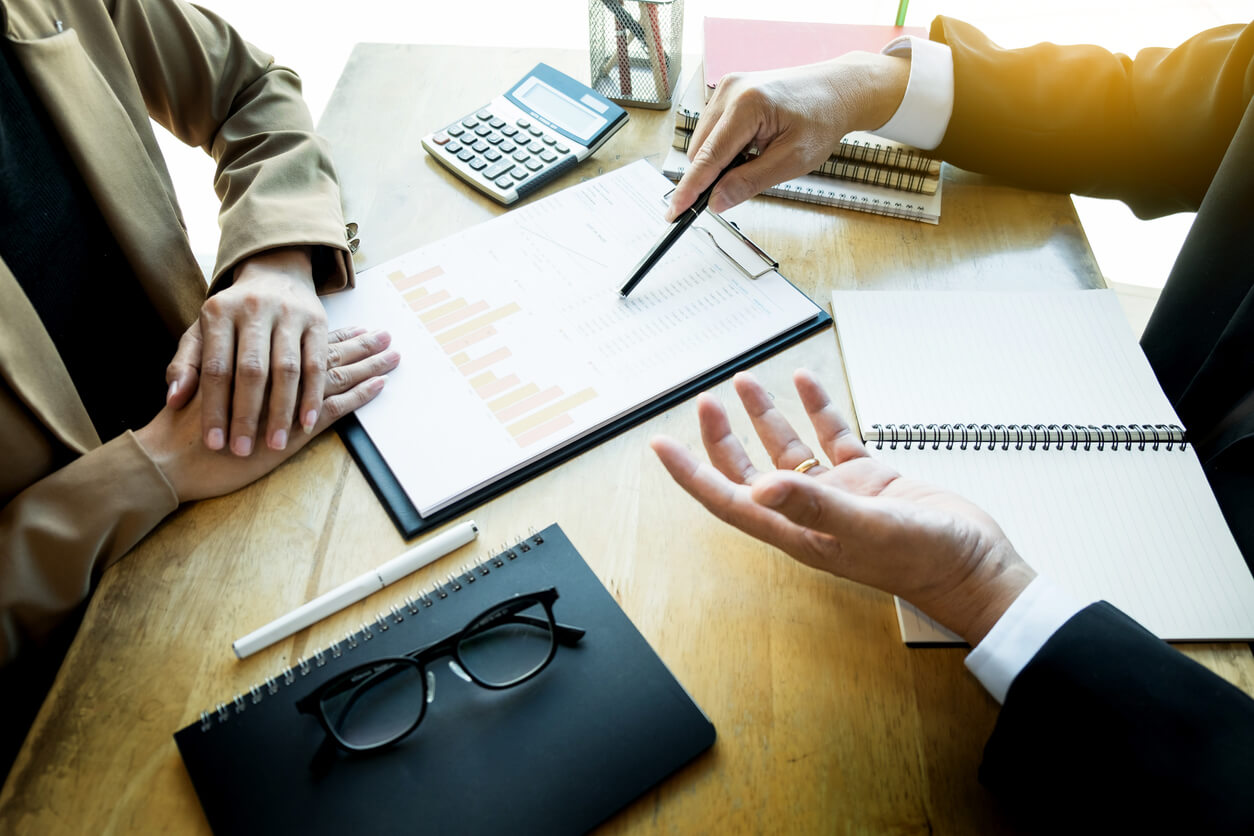 Two people discussing a financial report with charts at a desk with notebooks, glasses, and a calculator.