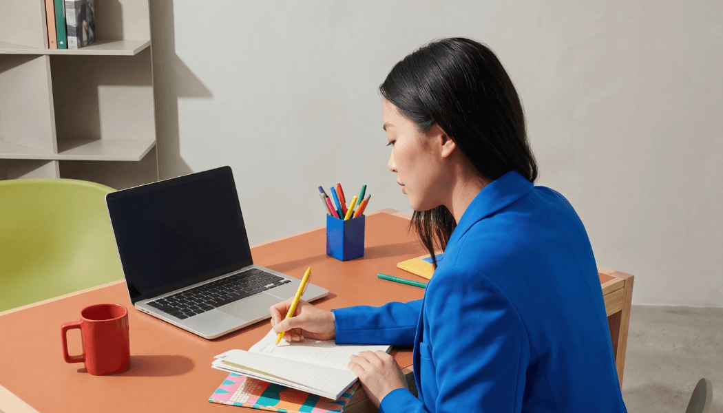 An assistant in a blue blazer writes with a yellow pencil in a notebook at a desk with a laptop, red mug, and colorful notebooks.