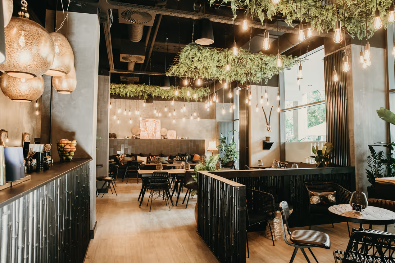 Modernes Café mit hängenden Glühbirnen, grünen Pflanzen an der Decke und gemütlichen Sitzbereichen mit Holzfußboden.