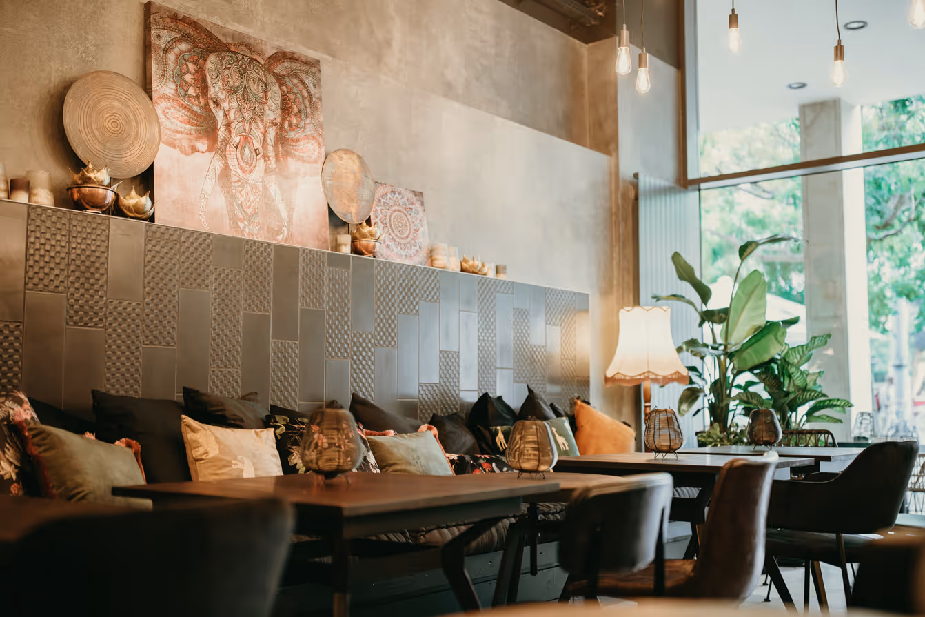 Gemütliche Café-Ecke mit gepolsterter Sitzbank, bunten Kissen, warmem Lampenlicht, Wanddekoration mit Elefantenmotiv und großen Pflanzen neben bodentiefen Fenstern.