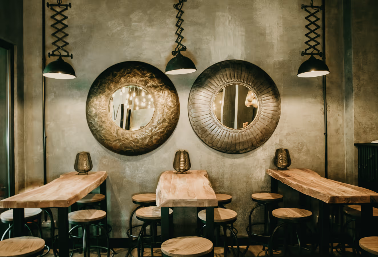 Rustikales Café mit Holztischen, hölzernen Hockern, zwei runden Metallspiegeln an der Wand und hängenden Pendelleuchten.