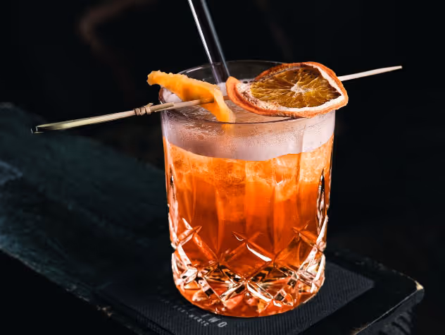 Ein orangefarbener Cocktail in einem facettierten Glas mit Eis, garniert mit einer getrockneten Orangenscheibe und einem Stück Orangenschale auf einem Holzspieß.