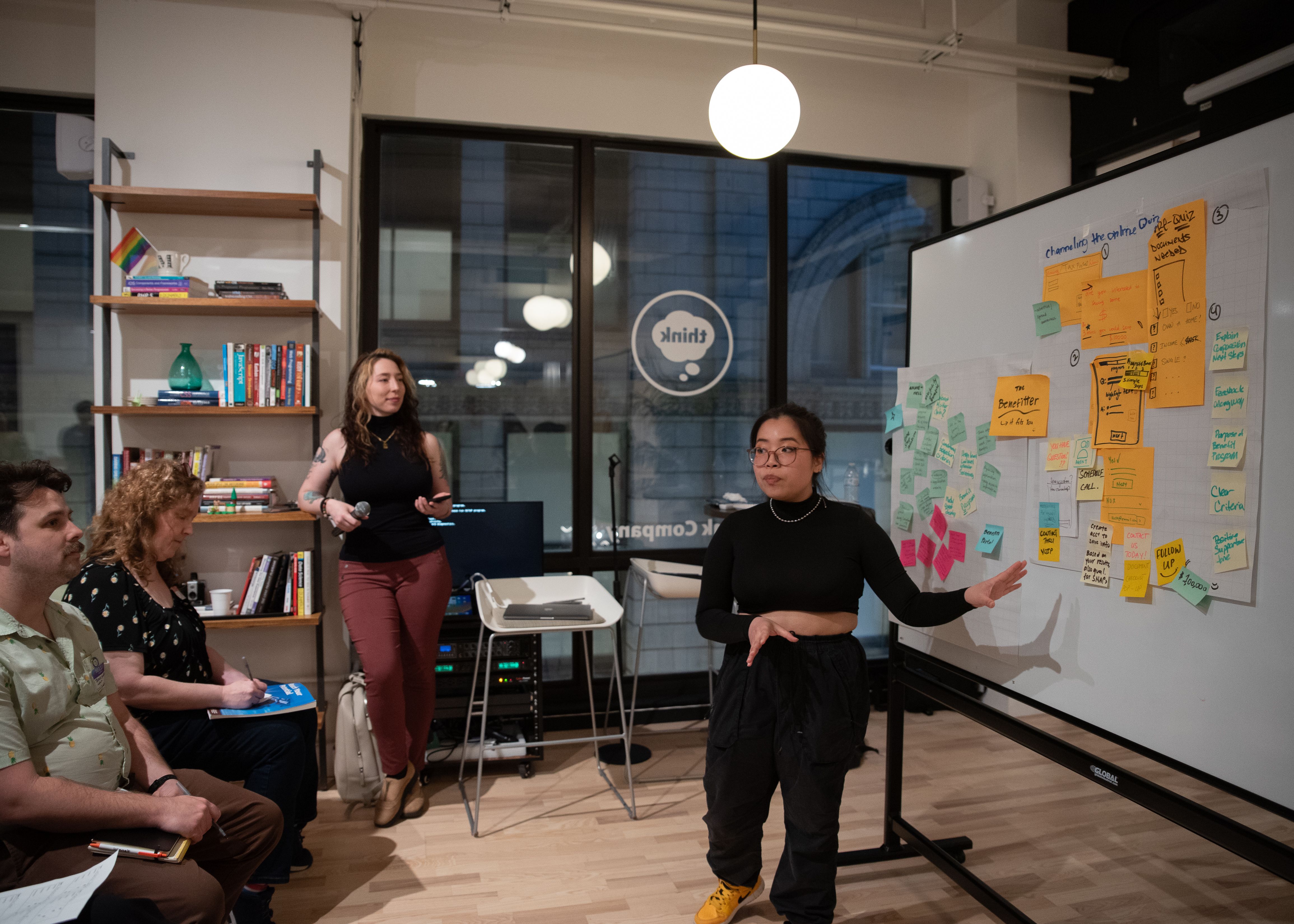 a design slam participant emphatically presenting her group project to the four judges, Jamie is mc-ing and smiling in the background as she watches attentively