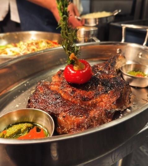Juicy bone-in ribeye steak served in a stainless steel pan with roasted pepper and rosemary at Duke's Steakhouse