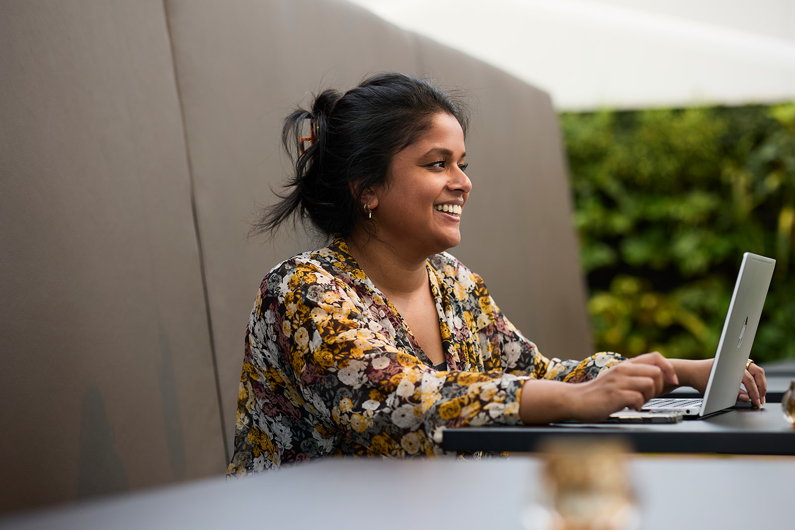 Smiling woman with dark hair in a floral blouse working on a laptop at a table.