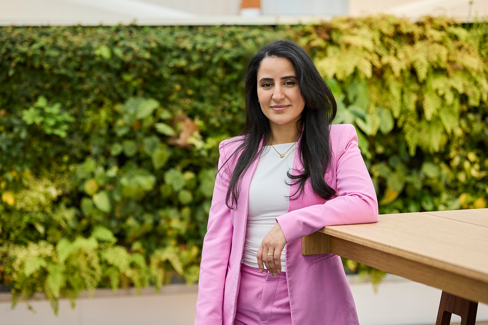 Woman with long dark hair wearing a pink suit and white top, leaning on a wooden table with a green leafy background.