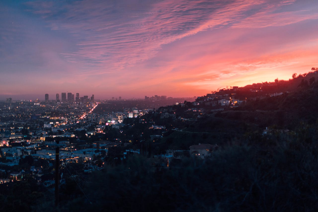 A cityscape at sunset with pink-hued clouds, city lights, and hillside homes.