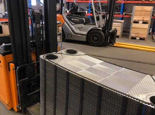 Gaffeltruck med orange gafler løfter en stor industriventilator i et lager med paller og en anden gaffeltruck i baggrunden.