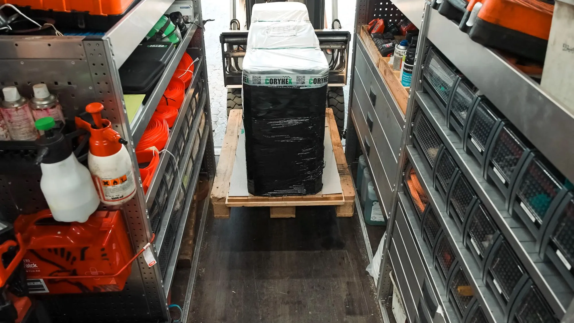 Gaffeltruck løfter en palle med en sortpakket kasse ind mellem værktøjsreoler i et lager.