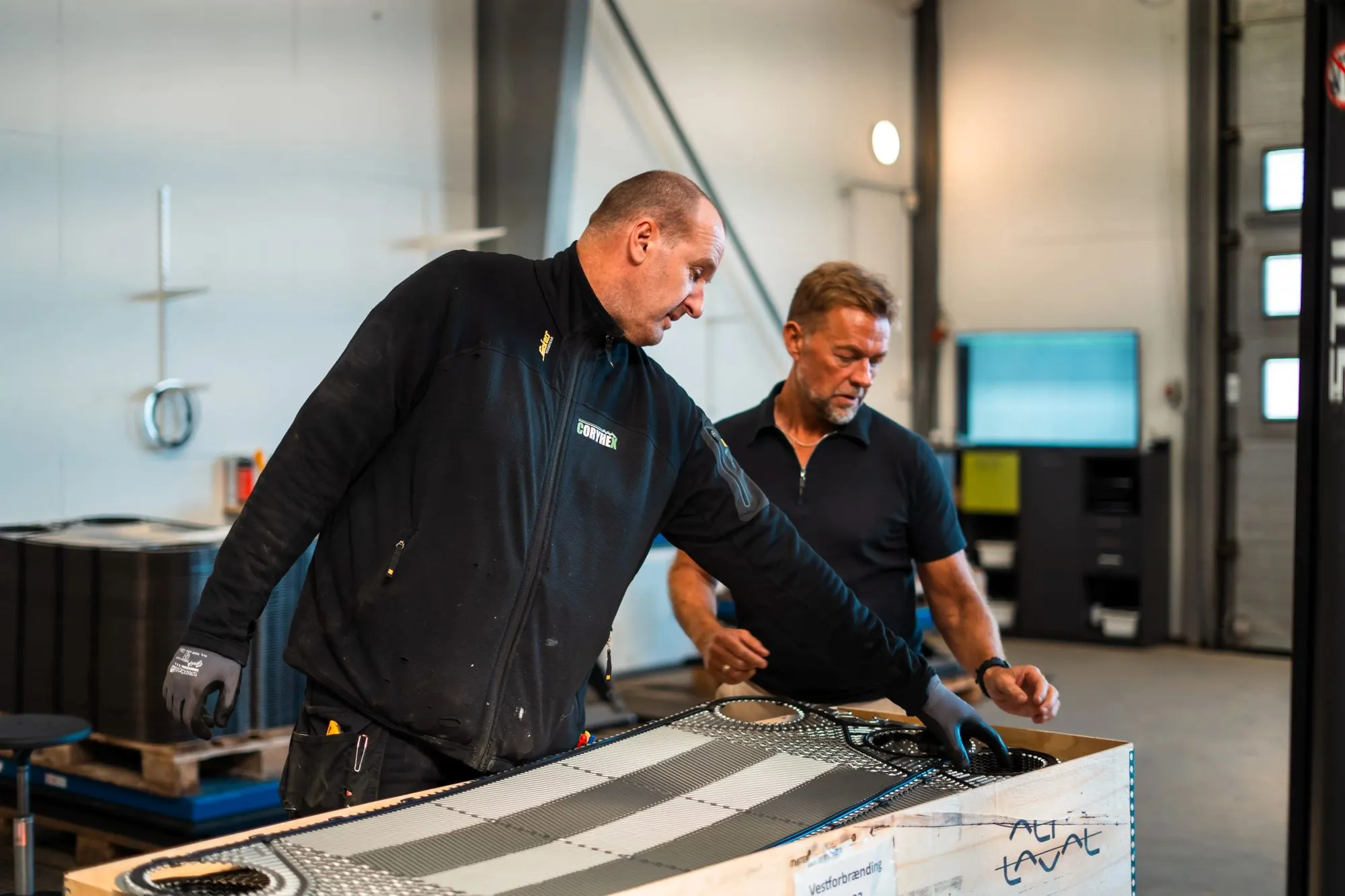 To mænd i arbejdsklæder arbejder sammen på et stort varmefordelingspanel i en industrihal.