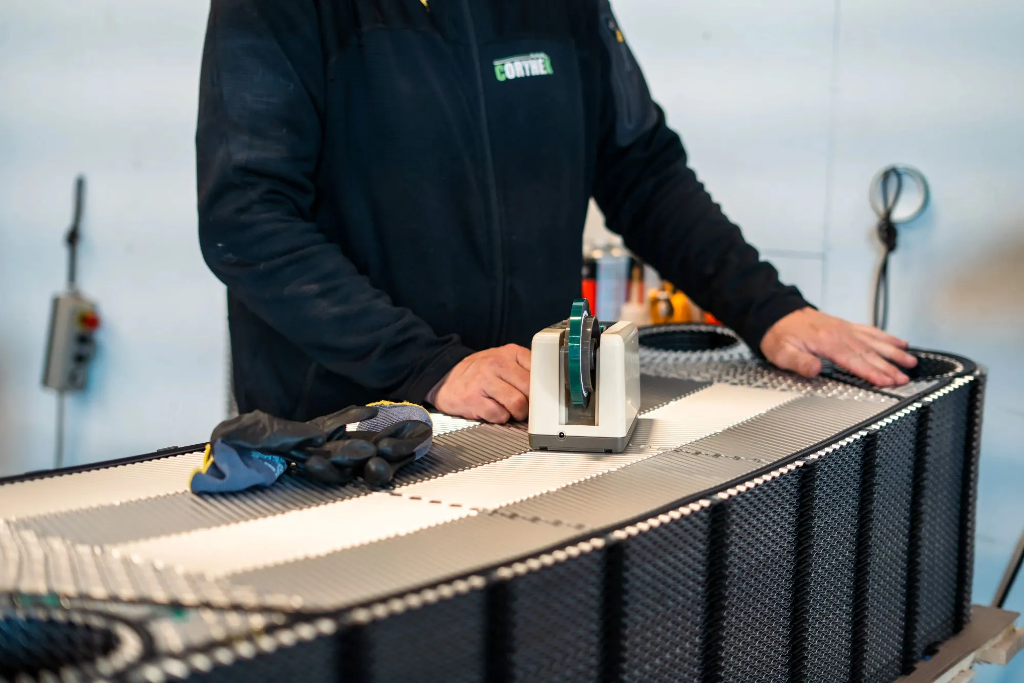 Person med mørk jakke hviler hænder på et industrielt sort- og hvidt bånd, hvor der ligger et par beskyttelseshandsker og en tape-afdispenser.