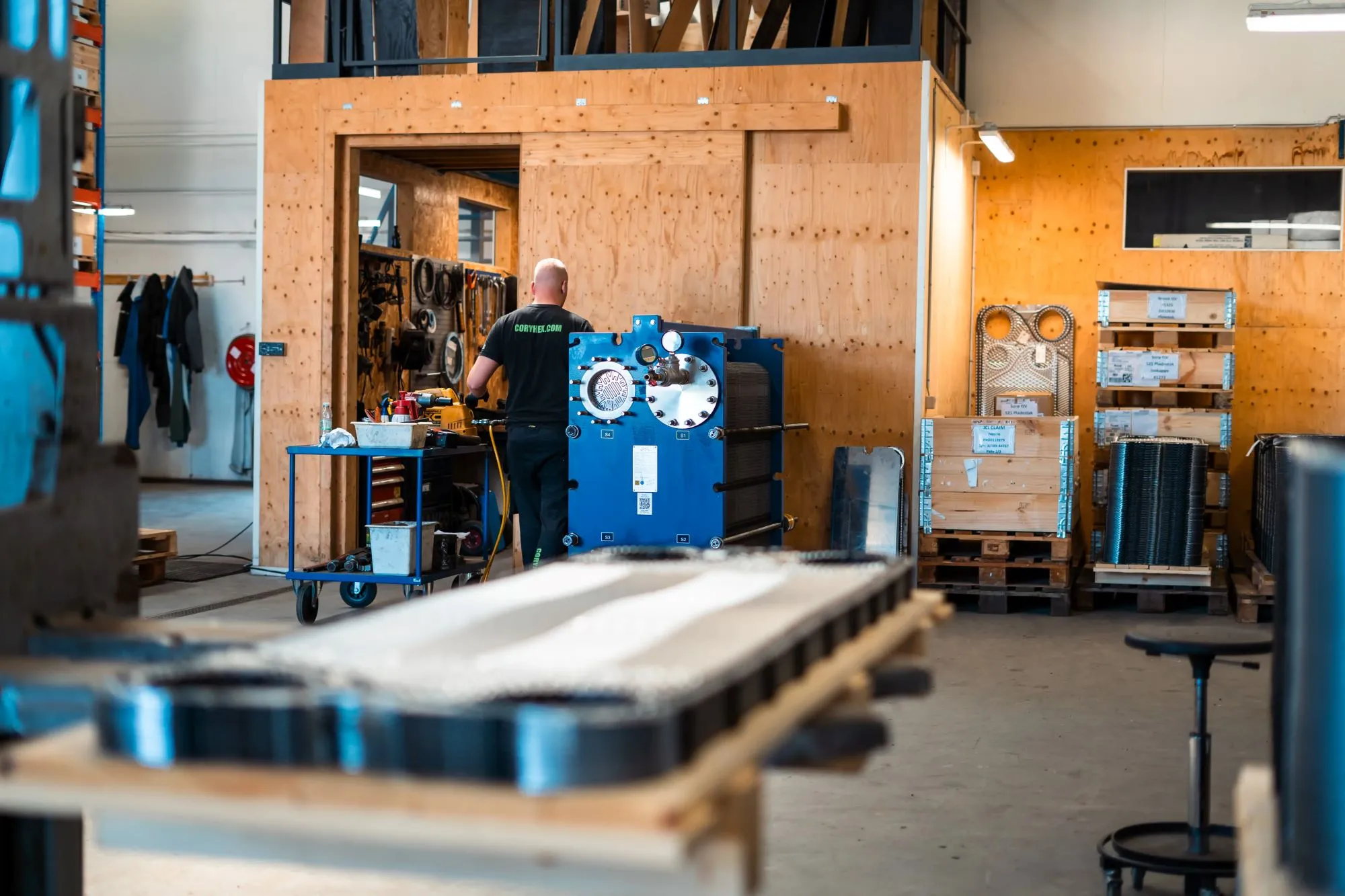 Mand i en fabrik eller værksted står foran et arbejdsbord med værktøj og en stor blå industri maskine mod en baggrund af trævægge og paller med udstyr.