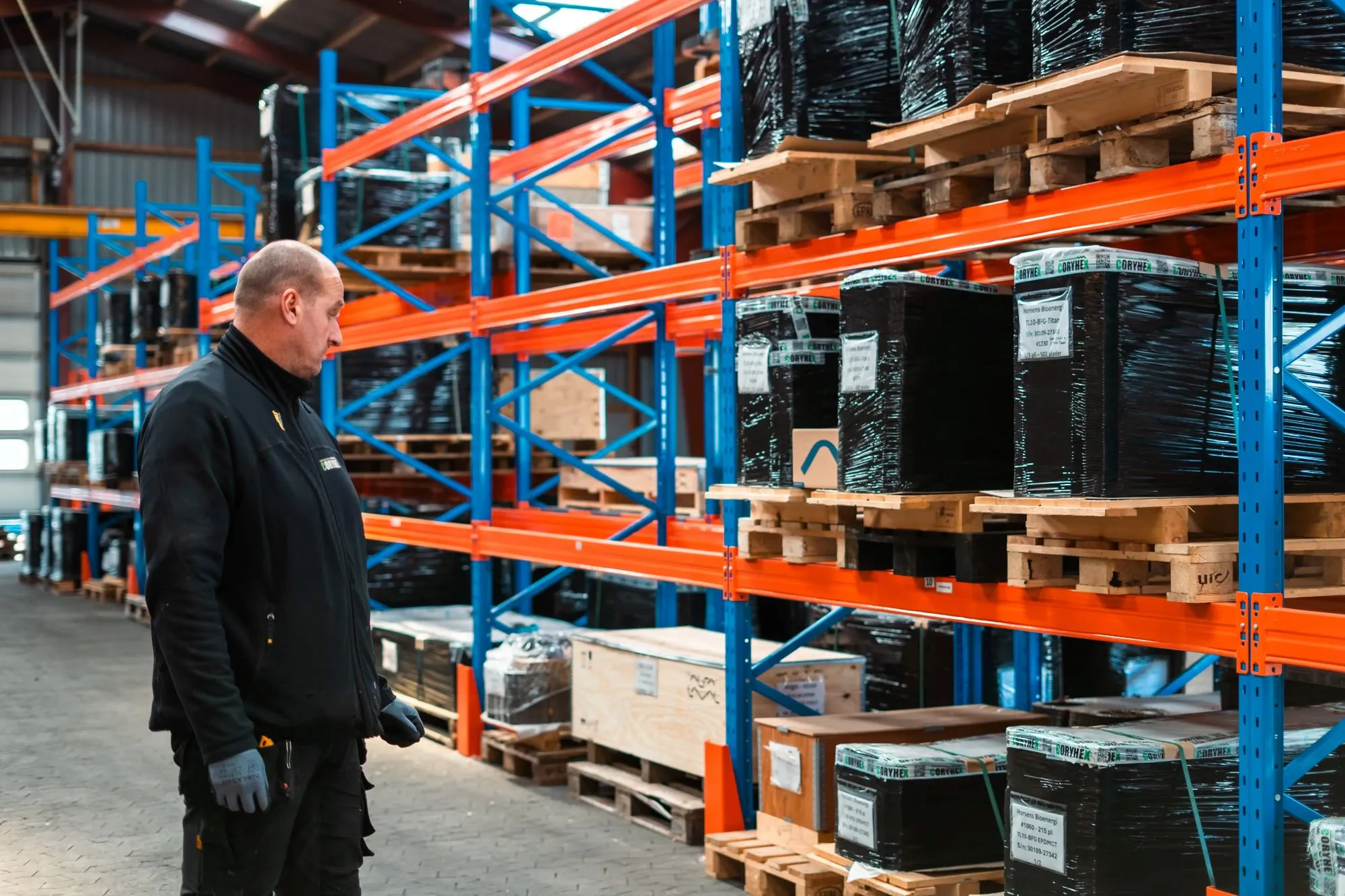 Mand i sort tøj står i et lager og kigger på stabler af paller pakket ind i sort plastik på orange og blå reoler.