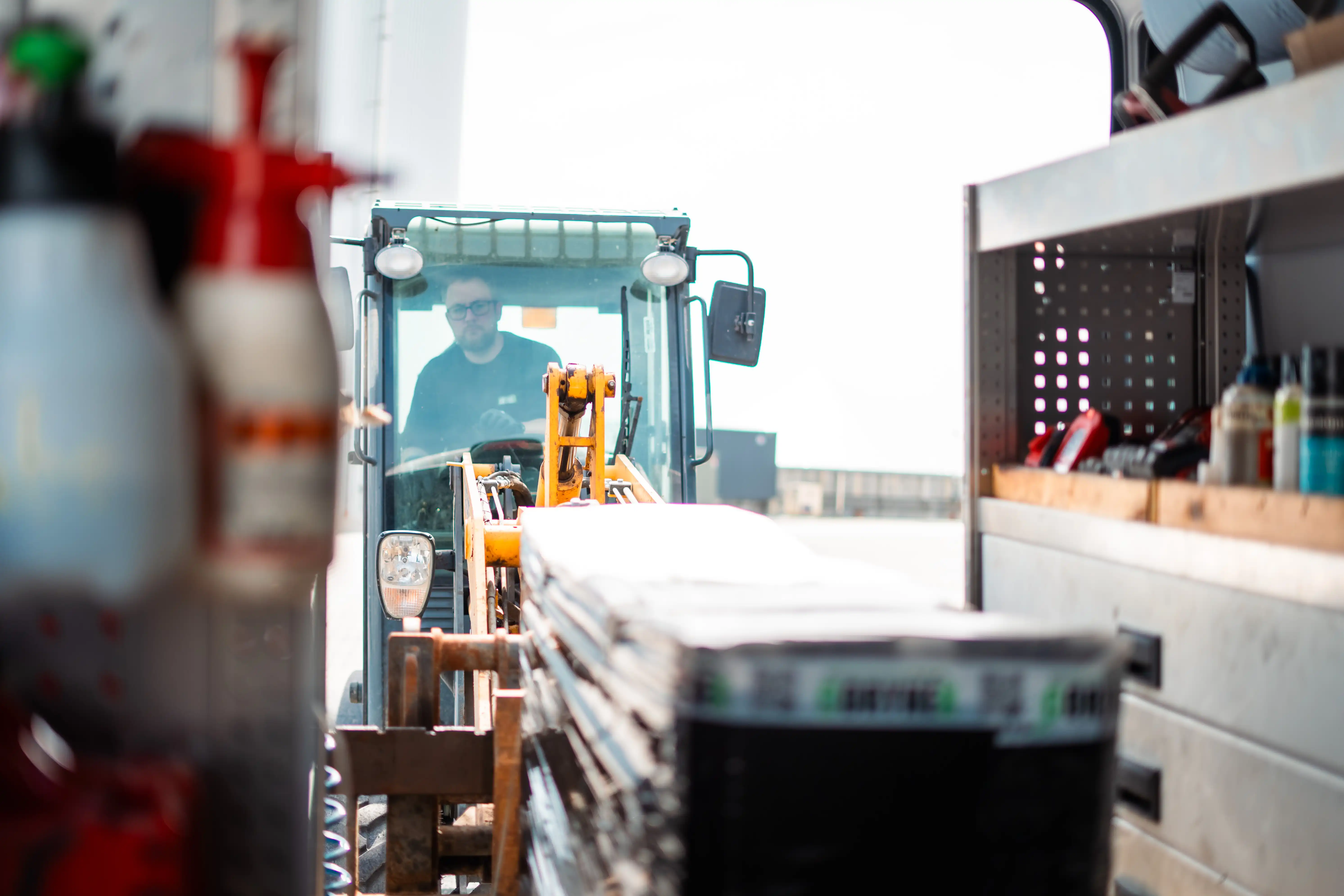 Mand, der kører en gaffeltruck, der løfter en palle med byggematerialer ved siden af en varebil med værktøj og udstyr.