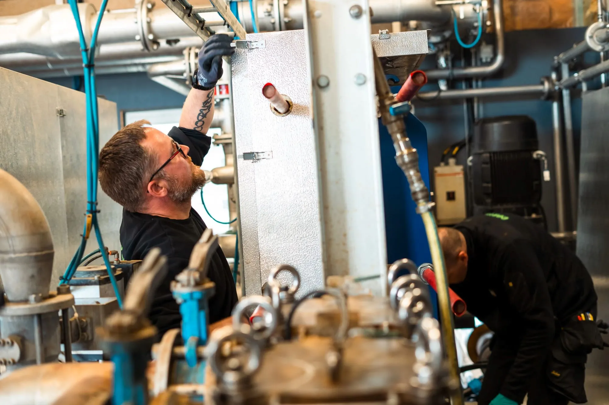 To mænd arbejder med industrimaskiner i et teknisk rum med rør og ventiler.