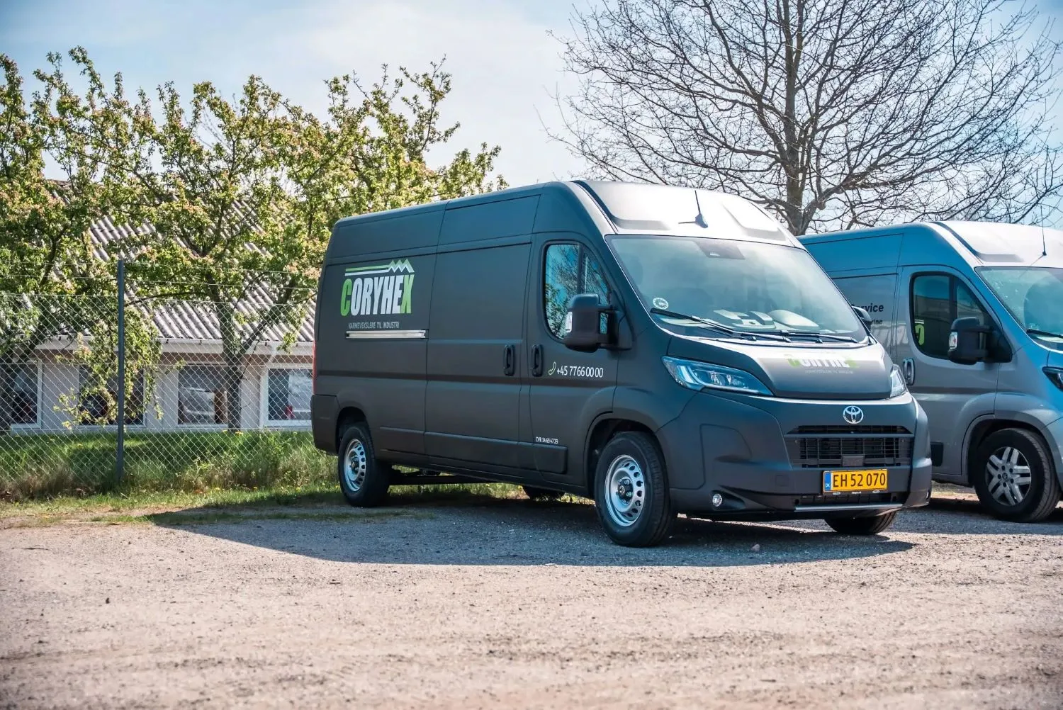 Sort virksomhedsvarevogn med Coryhex-logo parkeret på grusplads foran træer og hegn.