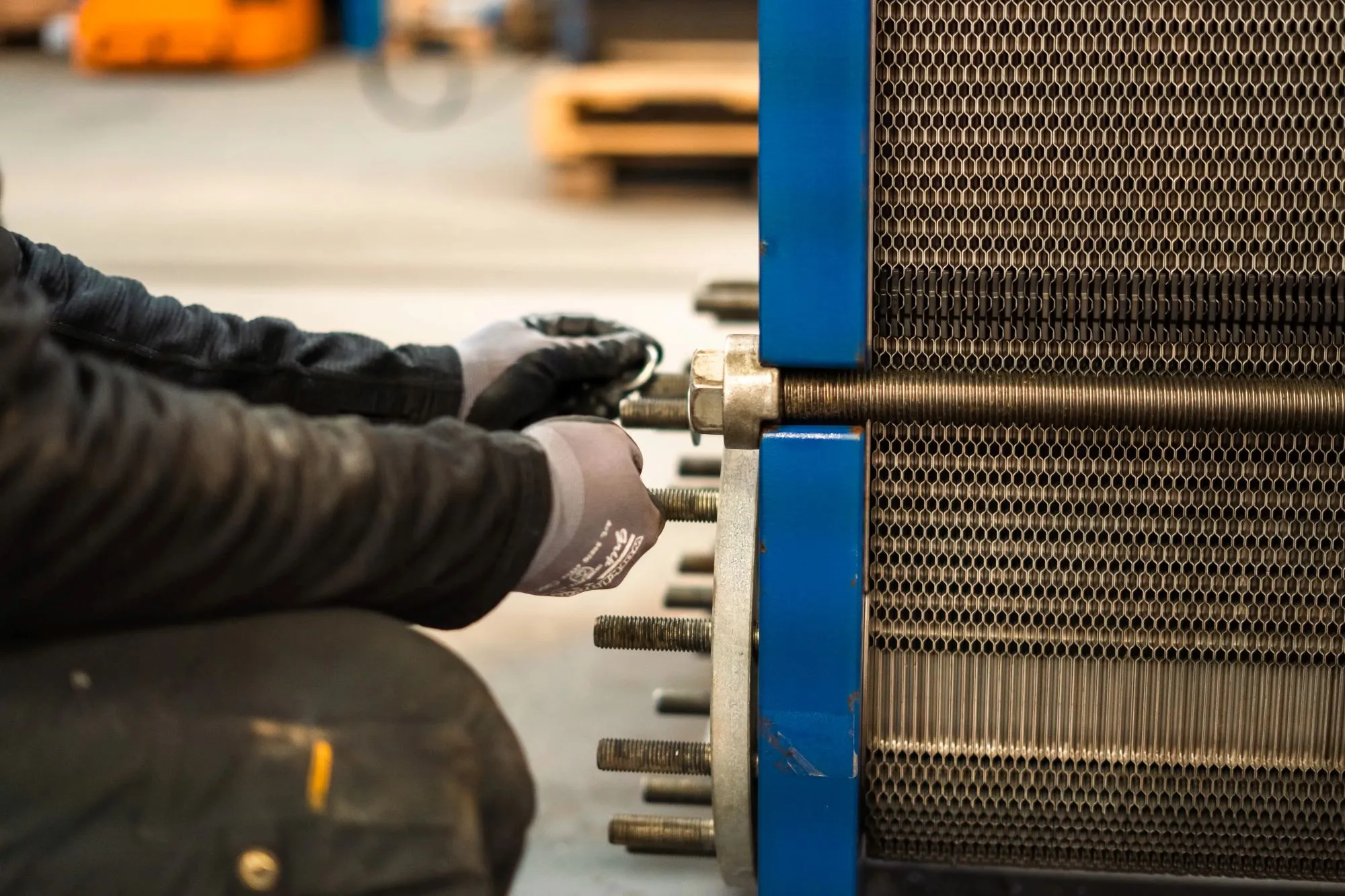 Nærbillede af arbejder med handsker, der justerer møtrik på en stor industriel varmeveksler med blå ramme.