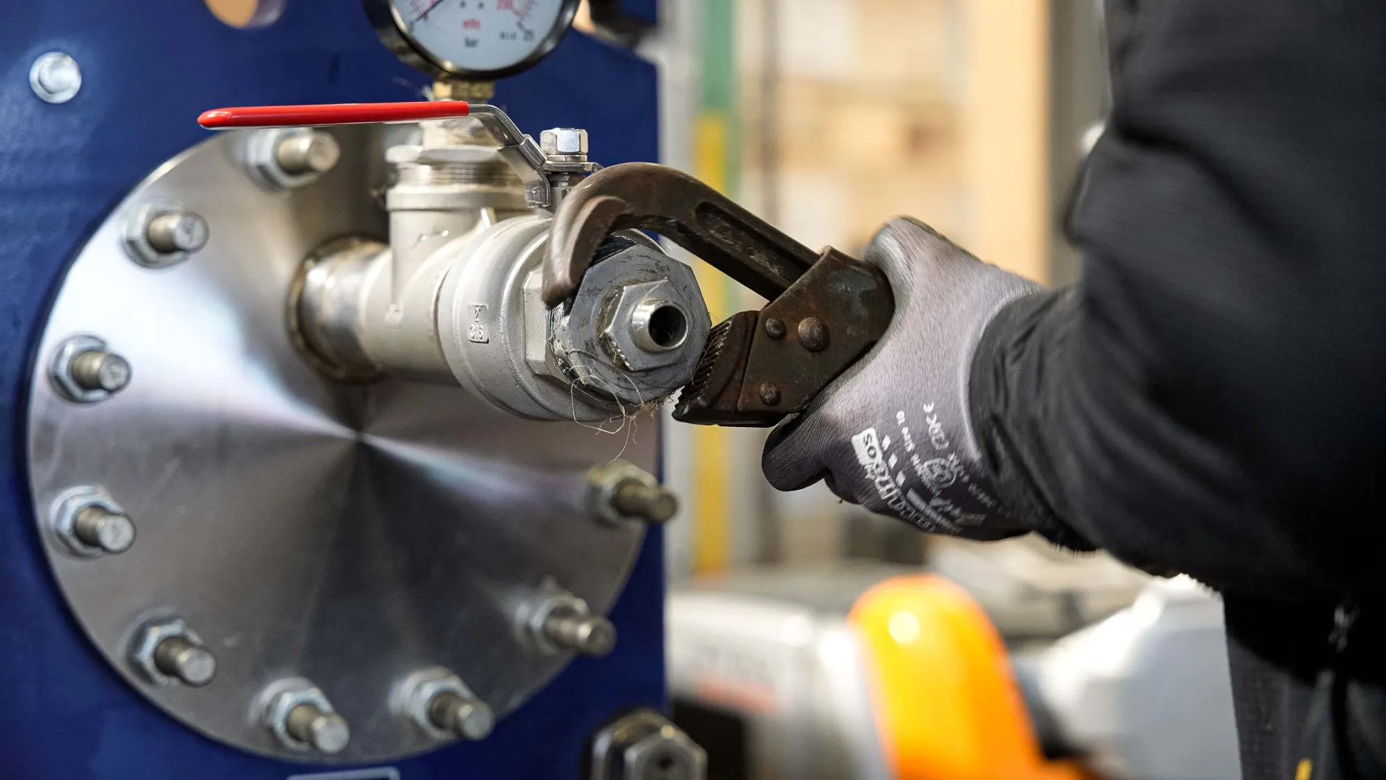 En tekniker iført grå sikkerhedshandsker bruger en skruenøgle til at justere en ventil på en industriel maskine.