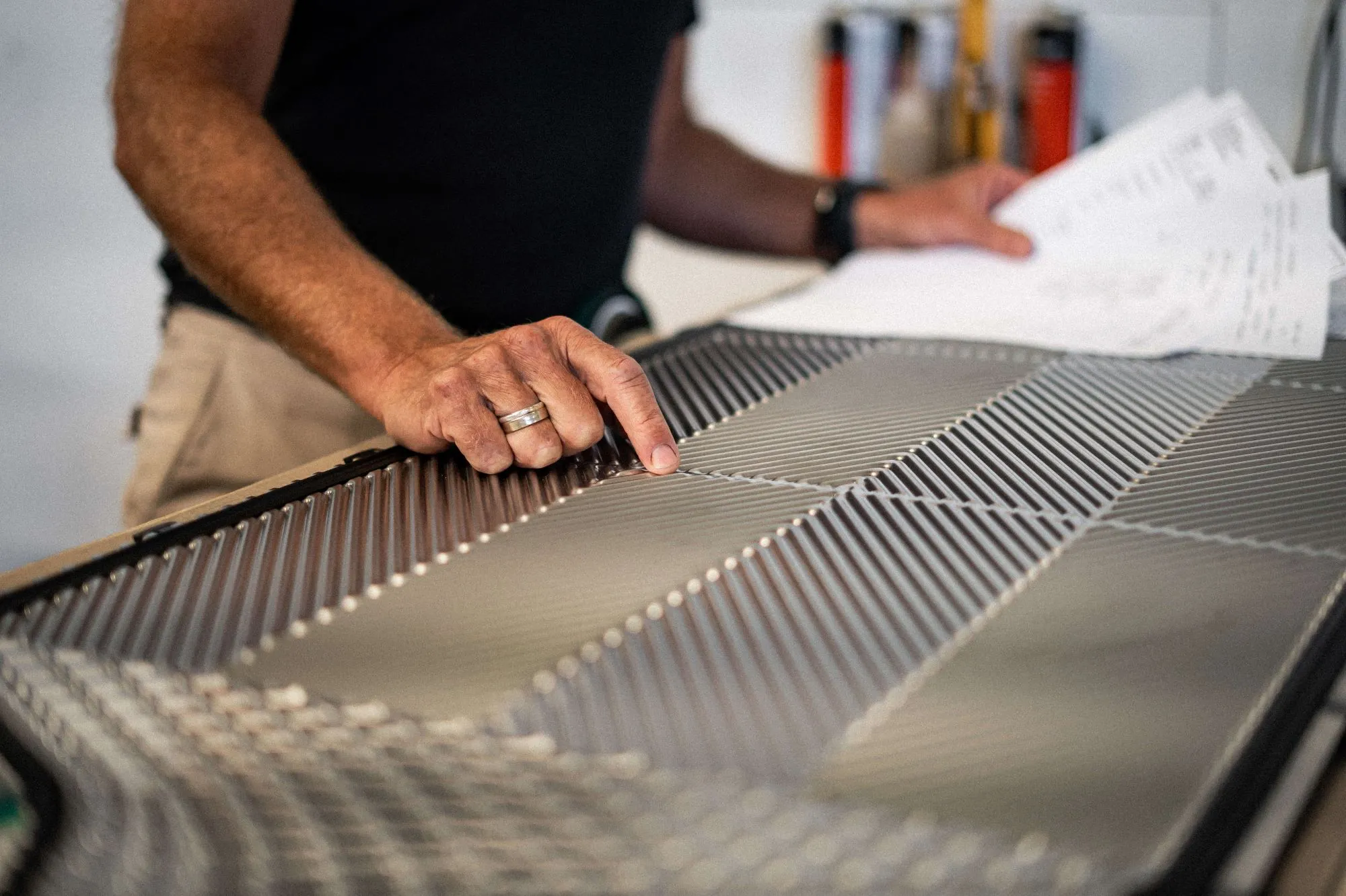 En person peger på et metalpanel med mønstret struktur og holder ark med skitser i den anden hånd.