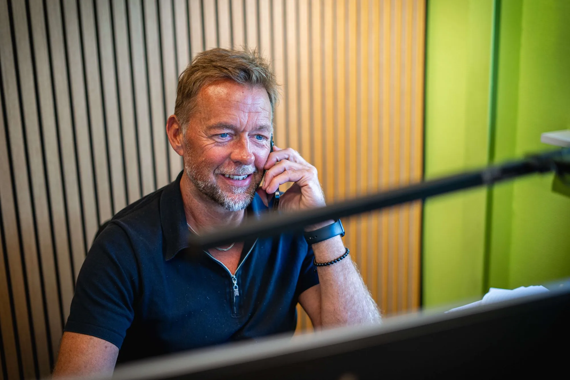 Mand med kort blondt hår og skæg taler i telefon foran en computer i et kontormiljø med træpaneler og grønne vægge.