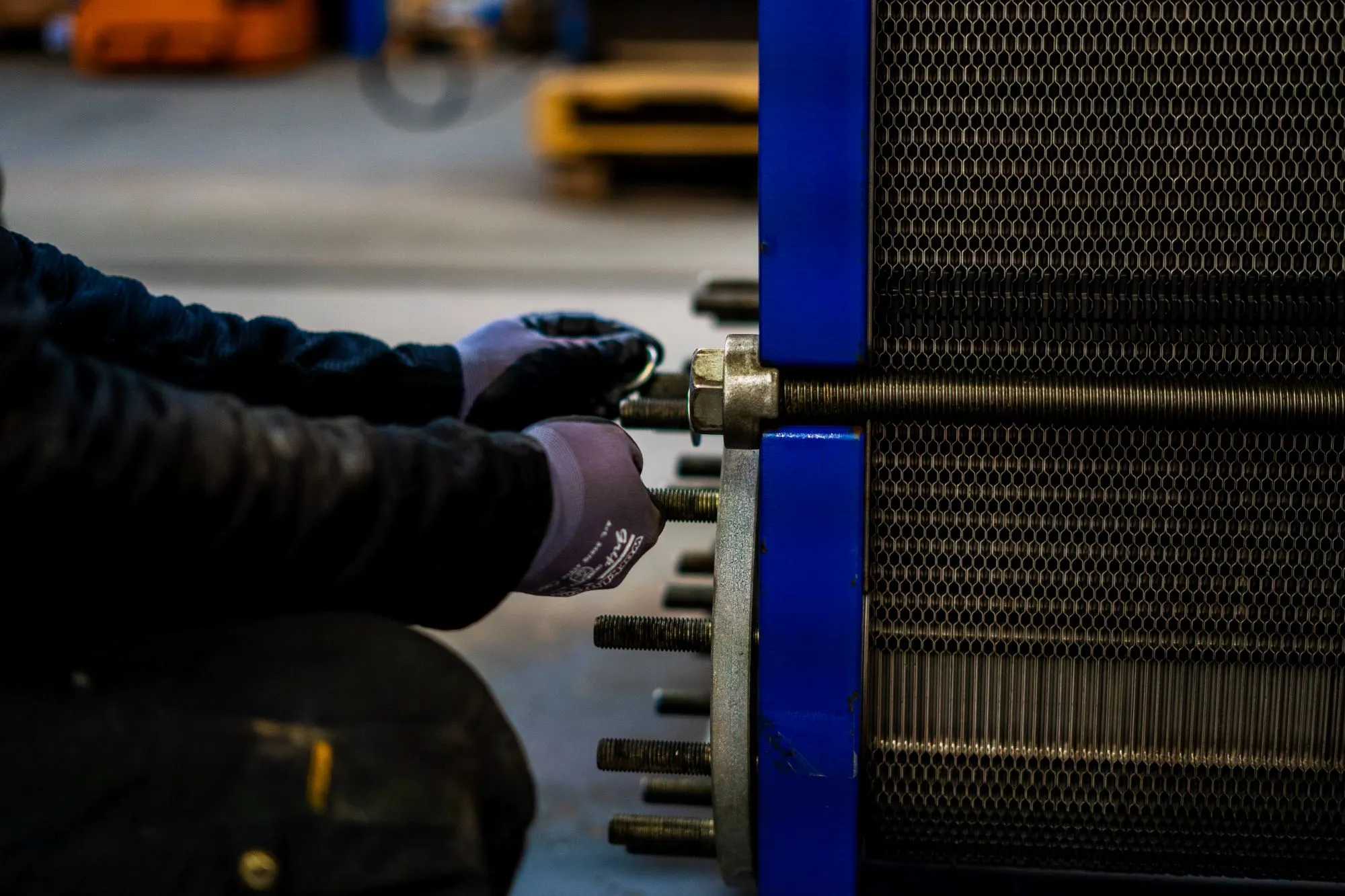 En person med handsker arbejder på at fastgøre bolte på en stor blå maskine med et metallisk gitter.