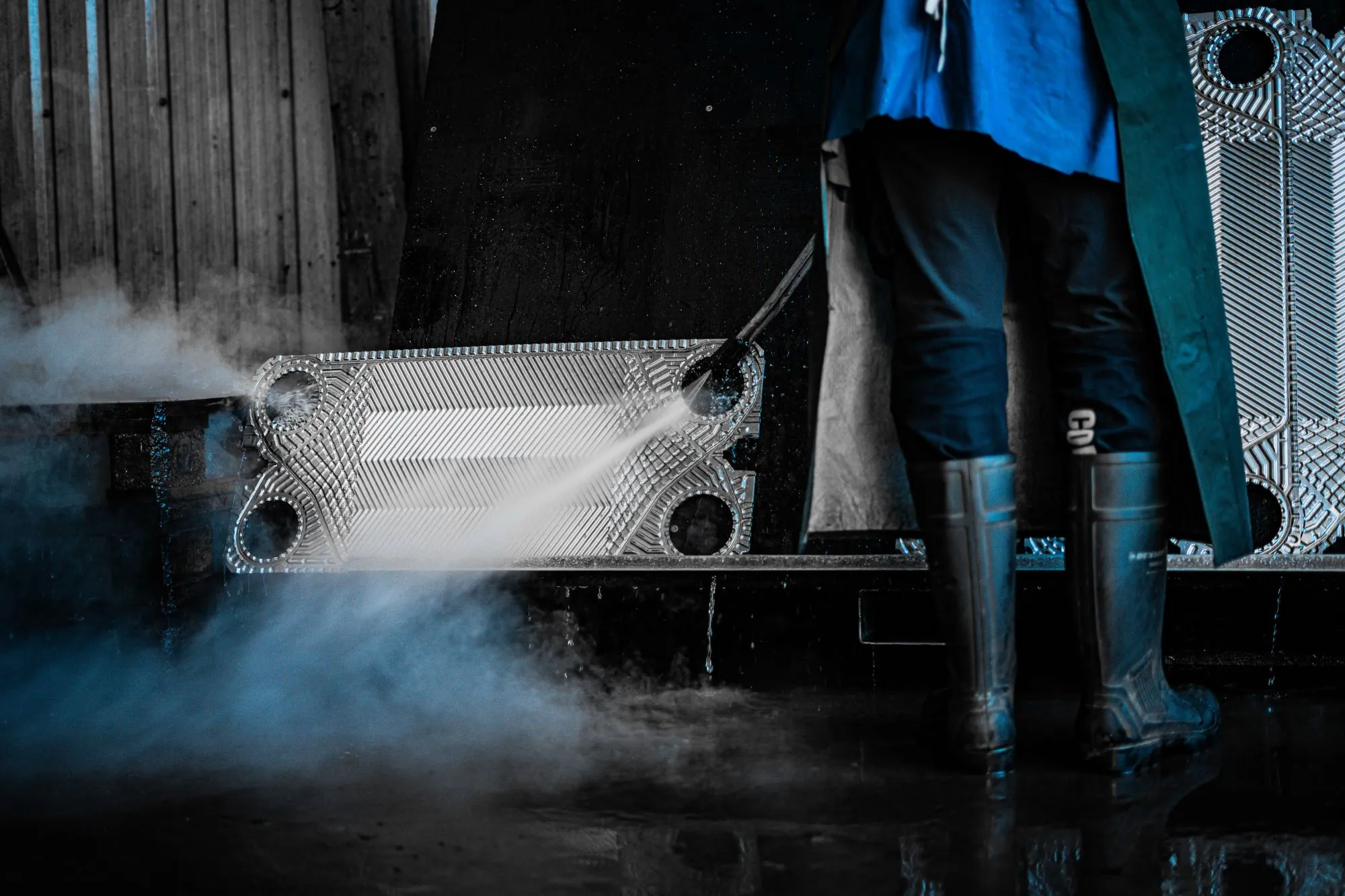 Person iført gummistøvler og blå tøj bruger en højtryksrenser til at rengøre metaldele i en industriel indstilling.