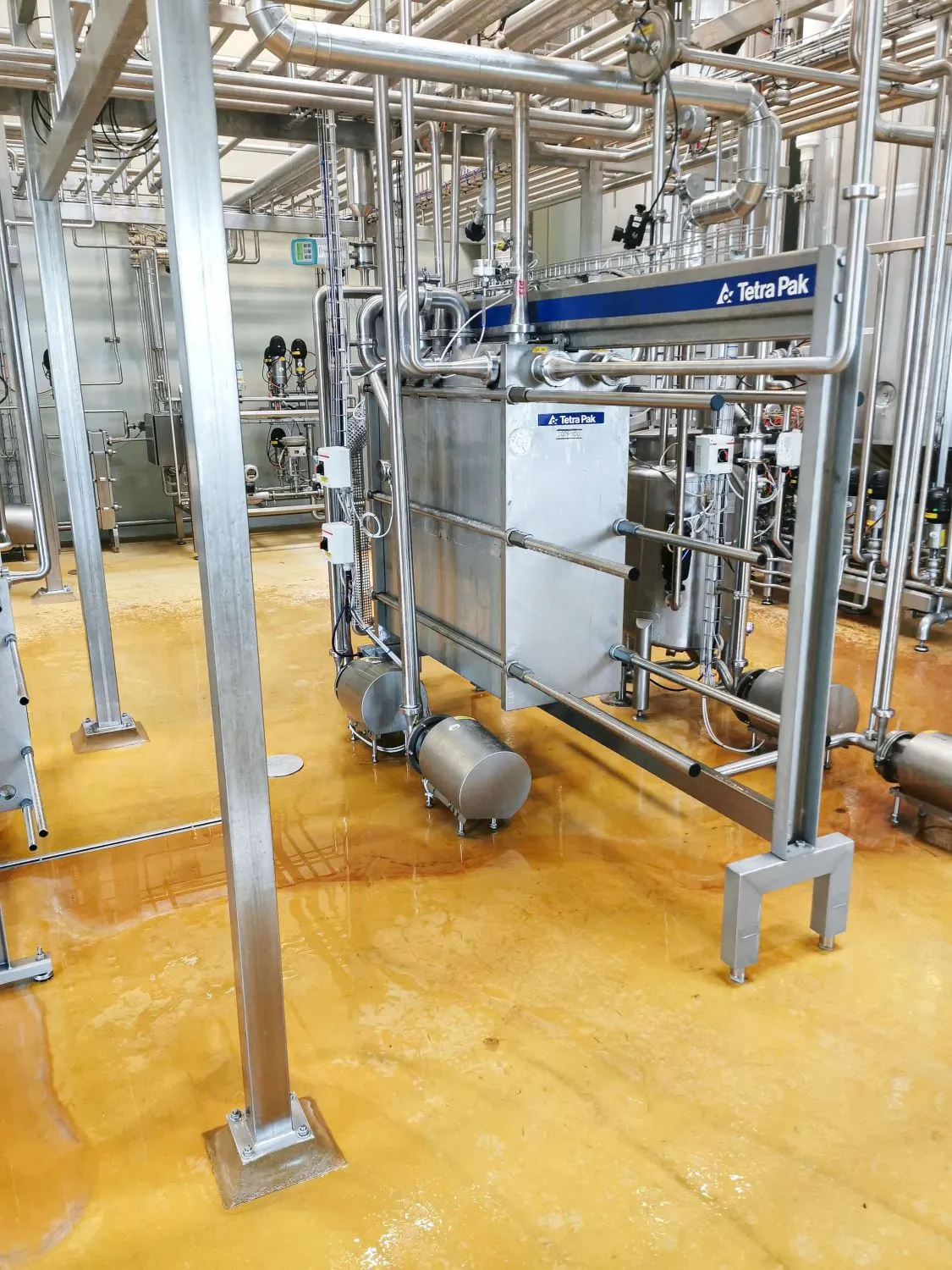 Rustfri stålindustrielt anlæg med Tetra Pak-mærket udstyr og kompleks rørføring på gulv med gul overflade.