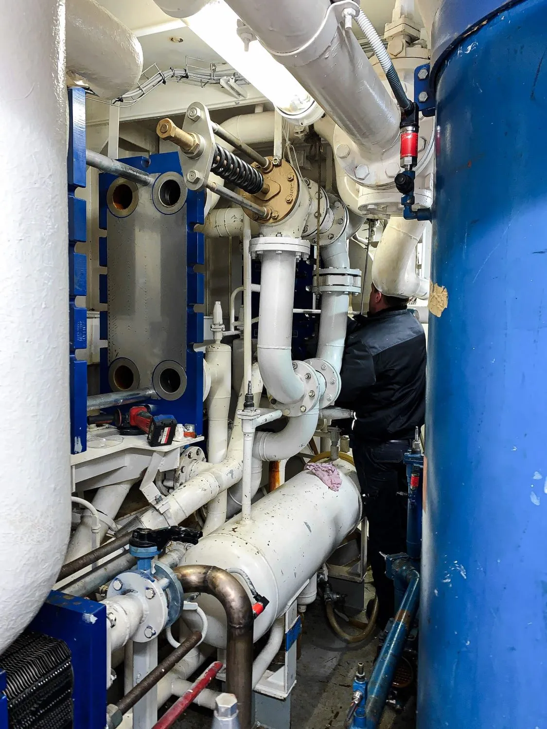 Tekniker arbejder med komplekse industrielle rørsystemer og maskineri med hvide og blå komponenter i et maskinrum.
