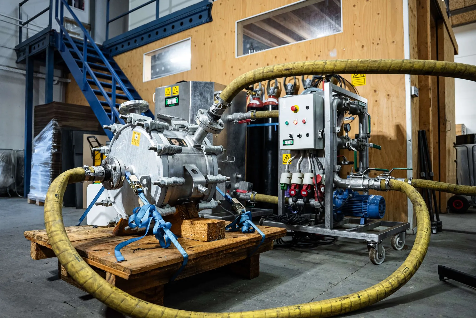 Industrielt udstyr med store gule slanger og metalrør i en lagerhal med trævæg og blå metaltrappe.