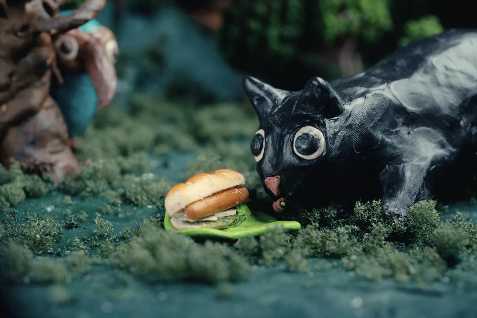 Stop-motion animation scene showing a handcrafted cat character interacting with a miniature sandwich prop.