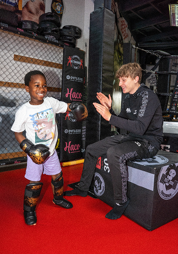 Jongen met bokshandschoenen en scheenbeschermers speelt boksen met coach in sportschool met rode vloer.