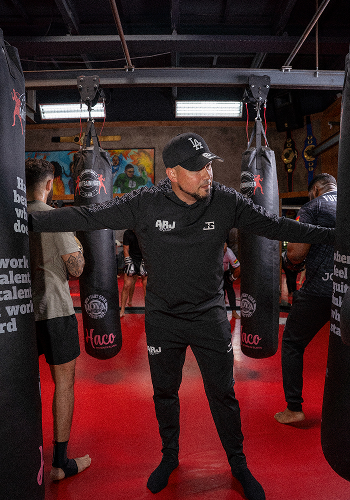 Persoon in zwarte sportkleding en pet staat tussen drie bokszakken in een sportschool met rode vloer.