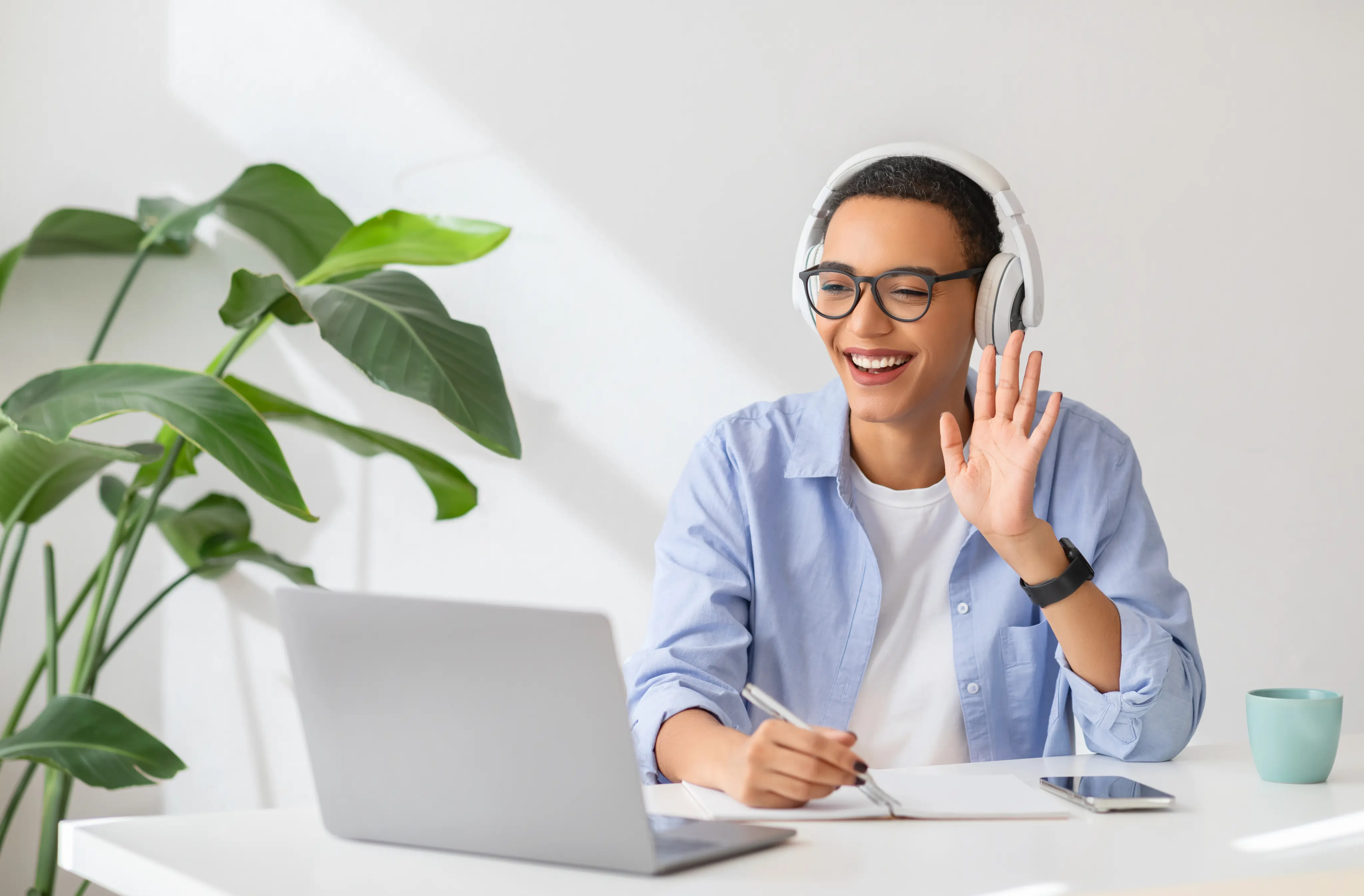 Junge Person mit Brille und Kopfhörern lächelt und winkt während eines Videoanrufs vor Laptop.