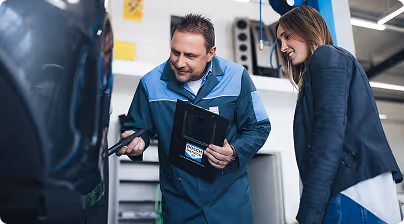 Mechanic showing a diagnostic tool to a female customer next to a car in a workshop.