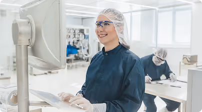Smiling lab technician in protective clothing looking at a computer screen in a clean laboratory setting.
