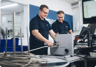 Two men in safety glasses working with industrial equipment and a computer in a modern workspace.