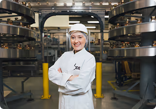 Smiling woman in white protective clothing and hairnet standing with arms crossed in an industrial factory setting with machinery.