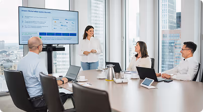 Businesswoman presenting data on a screen to three colleagues seated around a conference table with laptops in a modern office.