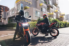 Two motorcyclists wearing helmets and jackets riding electric motorcycles on a city street with modern buildings in the background.