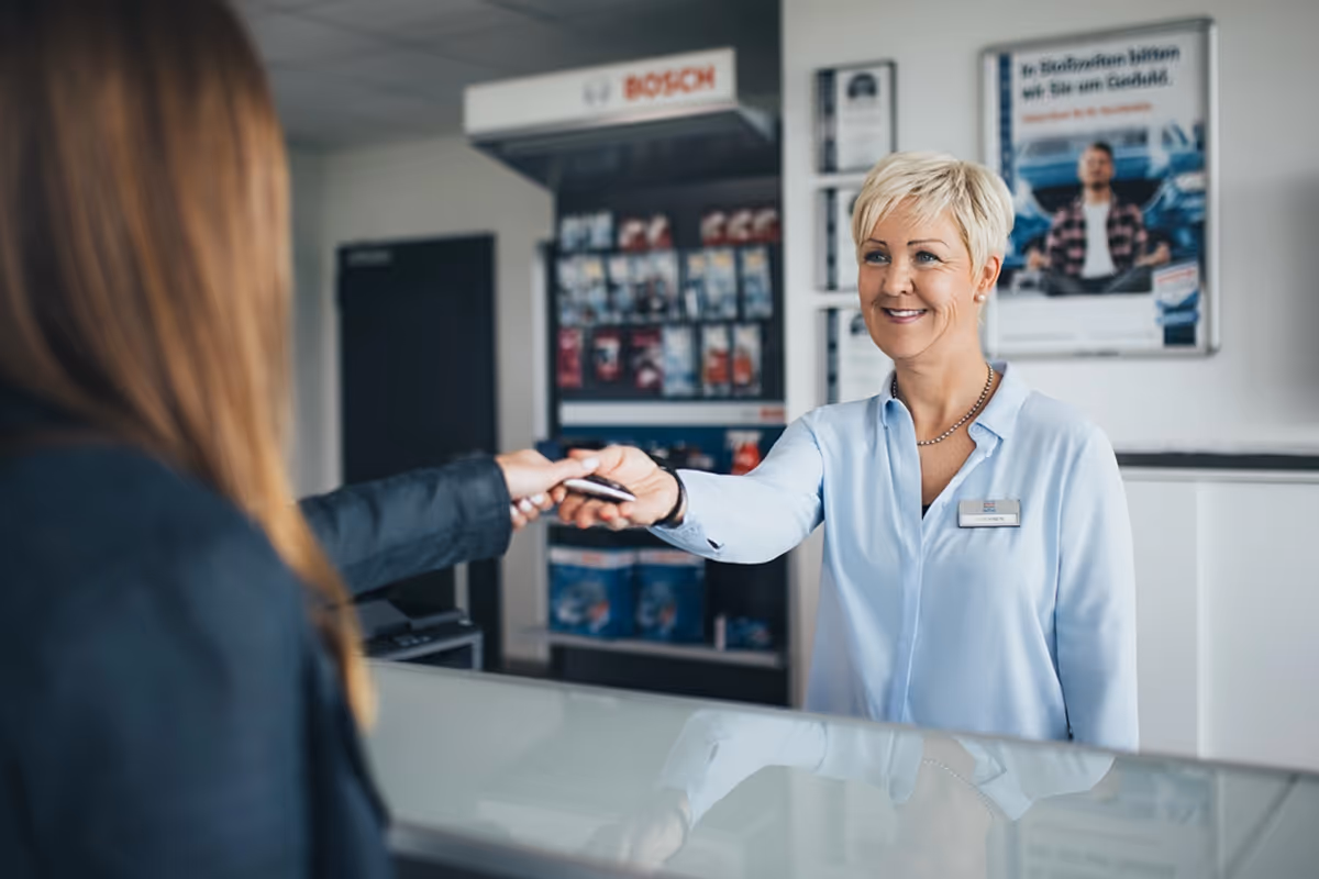 Smiling woman behind a counter hands a customer a card in a retail or service setting.