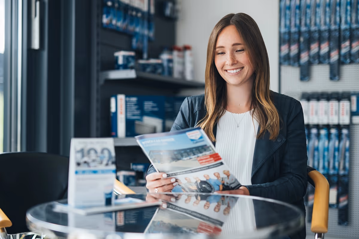 Smiling woman reading a brochure while seated at a glass table in a store or office.