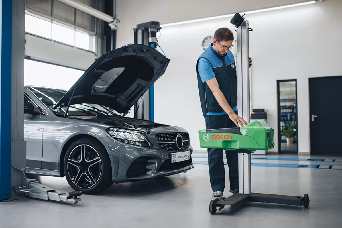Technician operating a Bosch automotive diagnostic machine next to a silver car with its hood open in a clean garage.