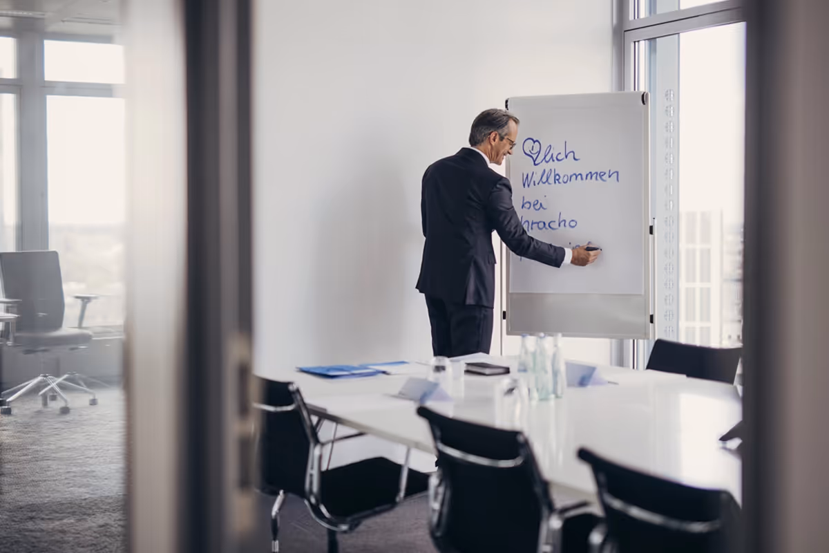 Man in a suit writing on a flipchart in a modern conference room with large windows.