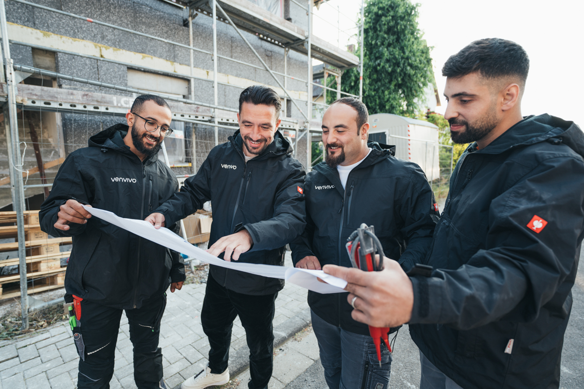 Four men in black Venvivo jackets discussing a blueprint at a construction site.