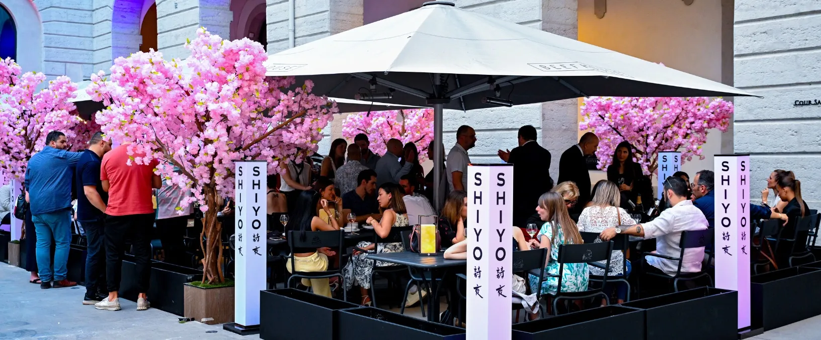 Terrasse de restaurant animée avec des groupes de personnes assises sous un grand parasol et entourées de cerisiers en fleurs artificiels roses. Des piliers décoratifs portant le nom 'SHIYO' sont également visibles, créant une ambiance festive et accueillante en extérieur.
