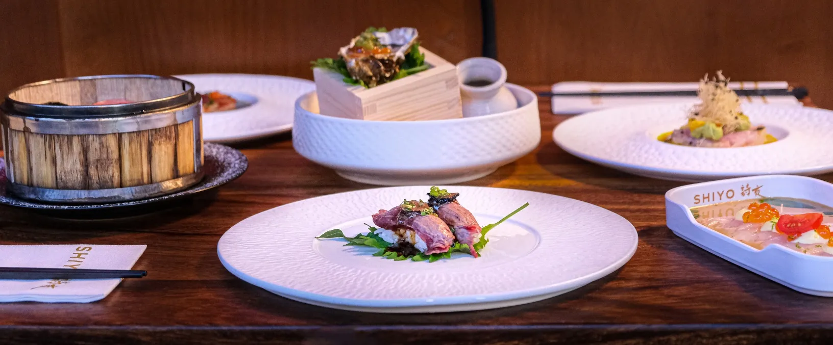 Une table en bois avec un assortiment de plats gastronomiques japonais : au premier plan, des tranches de viande sur un lit de riz et de feuilles vertes ; à l'arrière-plan, une huître sur de la glace, un cuiseur vapeur en bambou, et d'autres plats raffinés dans des assiettes blanches texturées.